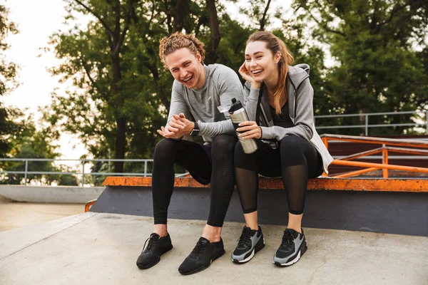 Photo of caucasian athletic couple guy and girl 20s in tracksuits laughing while sitting on parapet in green park after workout or jog on sunny summer day