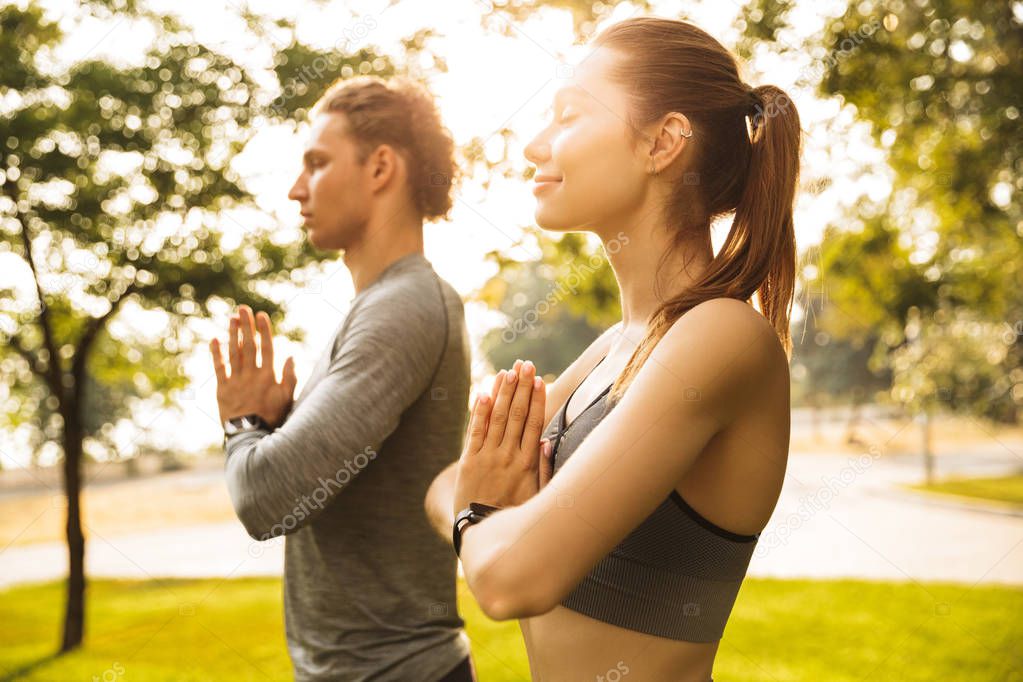 Image of athletic sporty couple man and woman 20s in tracksuits holding palms together while working out or doing yoga in green park