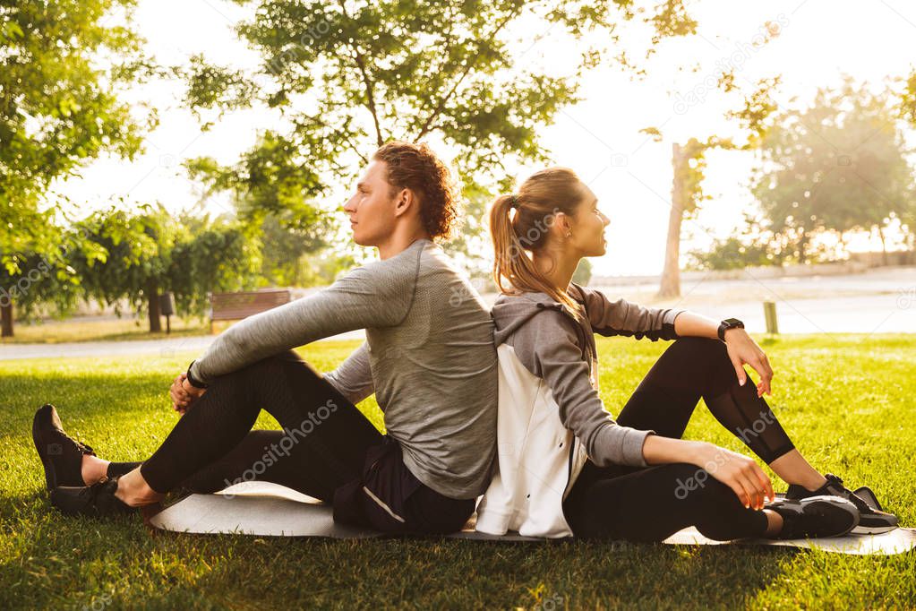 Image of young sporty couple man and woman 20s in tracksuits sitting on grass in green park back to back