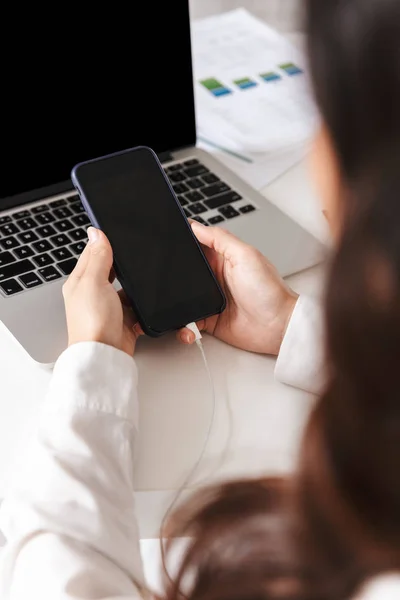 空白の画面携帯電話を保持しているラップトップ コンピューターを机の上に座っている白いシャツの女性のクローズ アップ — ストック写真