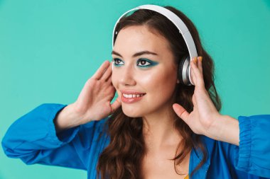 Görüntü closeup çekici kadın 20'li yaşlarda giyen yağmurluk ya da bir kenara seyir ve mavi arka plan üzerinde izole kablosuz kulaklık ile müzik dinlemek ceket