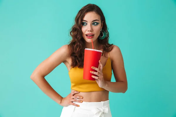 Photo of glamorous brunette woman 20s wearing fashion earrings drinking beverage or soda from big red paper cup and straw isolated over blue background