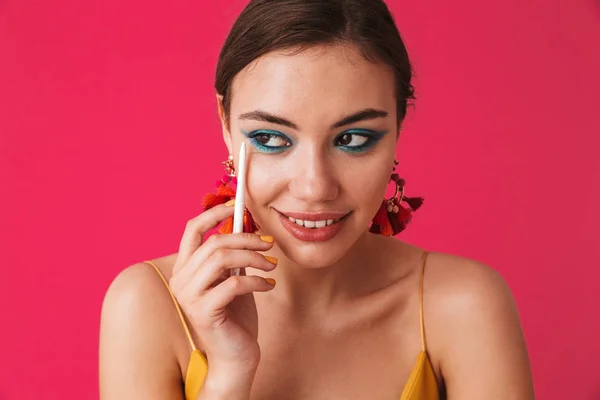 Photo of stylish trendy woman 20s wearing earrings looking aside and holding white pencil for makeup isolated over pink background