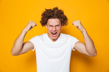 Güçlü kıvırcık 20'li yaşlarda erkekte çığlık ve sarı arka plan üzerinde izole kamera pazı gösterilen rahat t-shirt fotoğrafı