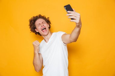 Cep telefonu sarı arka plan izole selfie çekerken yumruğunu kenetlenmesi kahverengi saçlı neşeli adam