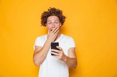 Sarı arka plan üzerinde izole smartphone tutarken casual beyaz t-shirt bağırarak şok veya sürpriz kıvırcık erkeklerde