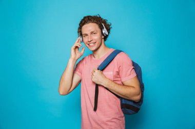 Mavi arka plan üzerinde mutlu gülümseyen adam 18-20 casual giyim ve kulaklık ile müzik dinlemek sırt çantası takmış kıvırcık saçlı portresi izole