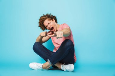 Bacaklar ile otururken kameraya dönük bir neşeli genç kıvırcık saçlı adam portresi mavi arka plan üzerinde çarpı işareti