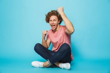 Bacaklar ile otururken kutluyor neşeli genç kıvırcık saçlı bir adam portresi mavi arka plan üzerinde çarpı işareti