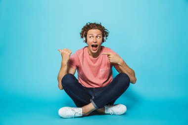 Portre mutlu bir genç kıvırcık saçlı adamın parmakları bacaklar ile otururken uzağa işaret kulaklık mavi arka plan üzerinde çarpı işareti