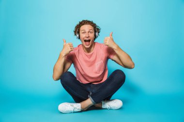 Portre mutlu bir genç kıvırcık saçlı adamın bacakları ile otururken başparmak gösterilmesini kulaklık mavi arka plan üzerinde çarpı işareti