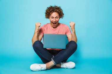 Fotoğraf başarılı adamın 20'li yaşlarda kahverengi kıvırcık saçlı gümüş laptop arıyorum ve bacaklar ile katta otururken sevindirici geçti, üzerinde mavi arka plan izole