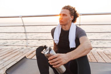 Açık havada kulaklık içme suyu ile müzik dinleme oturan plajda yakışıklı güçlü genç sporcunun fotoğrafı.