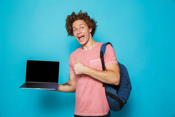 カジュアルな服や開いているノート パソコンと示す親指を保持分離青い背景上のバックパックを身に着けている巻き毛を持つ大学男の写真 — ストック写真