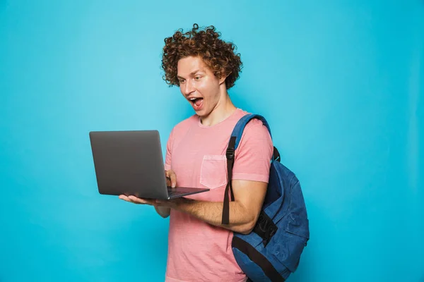 カジュアルな服と青い背景に分離されたオープンのラップトップを使用し 保持のバックパックを身に着けている巻き毛を持つ興奮して若い男のイメージ — ストック写真