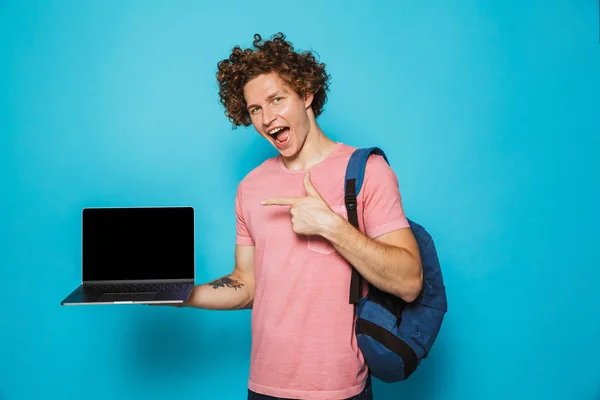 照片的大学男子卷曲的头发穿着休闲服装和背包持有打开笔记本电脑和指手指在屏幕上孤立的蓝色背景 — 图库照片
