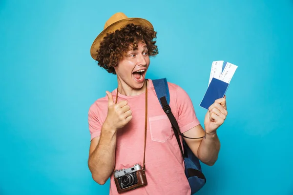 Photo of happy camera man 18-20 with curly hair wearing backpack and straw hat holding passport and tickets isolated over blue background