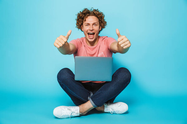Image of young joyful man 20s with brown curly hair smiling and showing thumbs up, while sitting on floor with legs crossed isolated over blue background