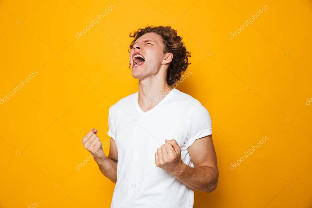 Photo of happy man 20s in casual t-shirt rejoicing and screaming with clenching fists isolated over yellow background