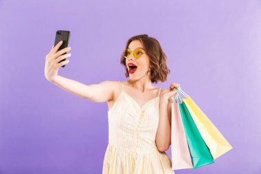 Sevimli güzel heyecanlı genç kadın fotoğraf tarafından cep telefonu üzerinde mor duvar arka plan alışveriş torbaları ile izole selfie olun.