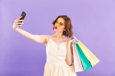 Sevimli güzel ve genç birbayan fotoğraf tarafından cep telefonu üzerinde mor duvar arka plan alışveriş torbaları tutan izole selfie olun.