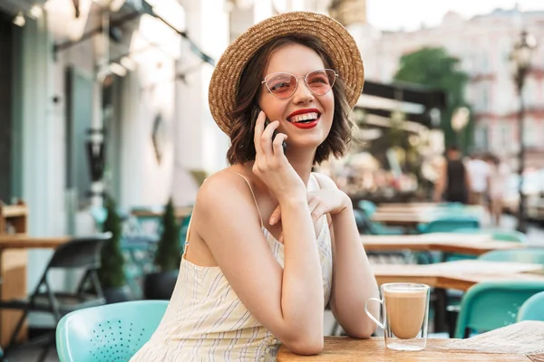 Joyful woman in dress and straw hat talking by smartphone while sitting by the table in cafe
