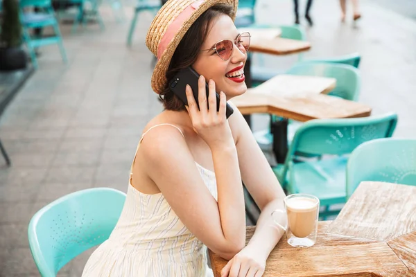 Side view of Happy woman in dress and straw hat talking by smartphone while sitting by the table in city cafe