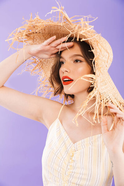 Close up portrait of a pretty young woman dressed in dress and straw hat looking away over violet background