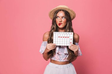 Portrait of brunette beautiful woman 20s wearing straw hat holding pms calendar isolated over pink background in studio clipart