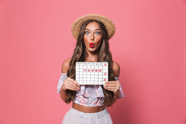 Портрет изумленной европейской женщины 20 лет, носящей соломенную шляпу, держащей велосипедный календарь, изолированный на розовом фоне в студии

