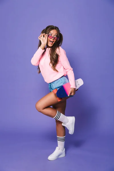紫色の背景に分離されたスケート ボードでポーズのスエット シャツで素敵な女の子の完全な長さの肖像画 — ストック写真