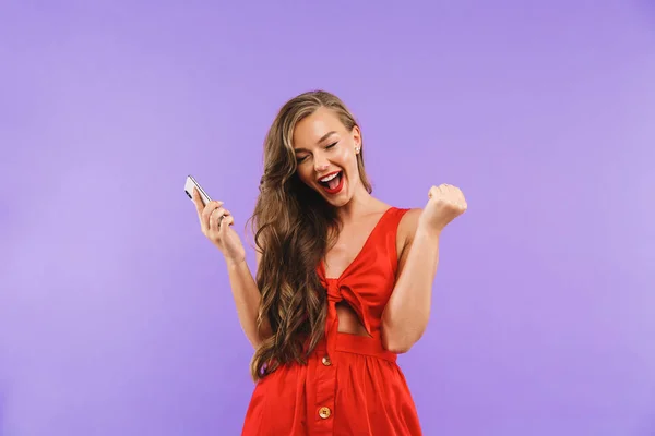 图像特写的快乐年轻女子20S 穿着红色礼服尖叫和喜悦 而持有手机站在紫色背景隔离 — 图库照片