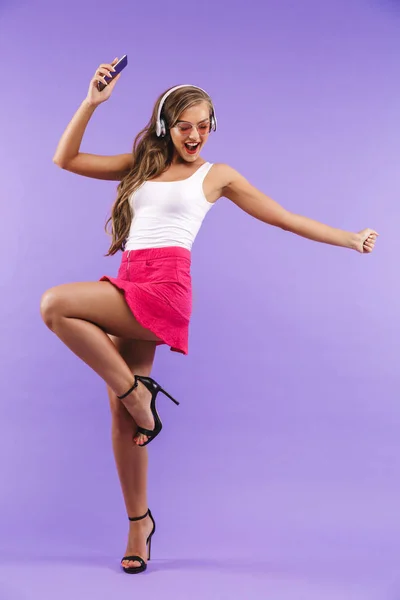 全长画像的快乐精力充沛的女人20S 在夏天的衣服和太阳镜跳舞 而听音乐通过无线耳机隔离紫色背景 — 图库照片