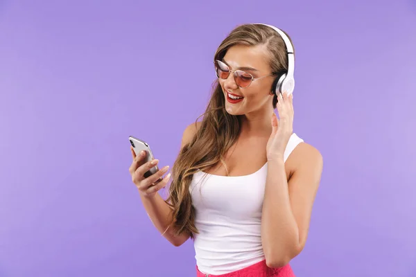 美丽的女人的肖像20S 在夏天的衣服和太阳镜听音乐时 使用无线耳机和智能手机隔离紫色背景 — 图库照片