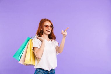 Alışveriş torbaları, uzağa işaret taşıyan bir gülümseyen genç kız menekşe arka plan üzerinde izole duran güneş gözlüğü portresi