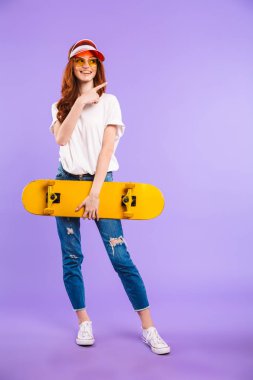 Neşeli bir genç kız kaykay, uzağa işaret tutan şapka ve güneş gözlüğü izole mor arka plan üzerinde duran tam uzunlukta portresi