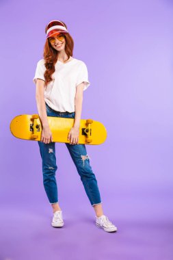 Tam uzunlukta portre kaykay holding neşeli bir genç kızın şapka ve güneş gözlüğü izole mor arka plan üzerinde duran