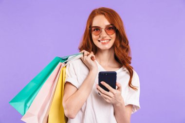 Cep telefonu kullaranarak alışveriş çantaları taşırken bir gülümseyen genç kız menekşe arka plan üzerinde izole duran güneş gözlüğü portresi