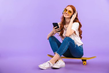 Cep telefonu, kulaklık ile müzik dinlemek tutan bir gülümseyen genç kız menekşe arka plan üzerinde izole bir kaykay üzerinde oturan güneş gözlüğü tam uzunlukta portresi