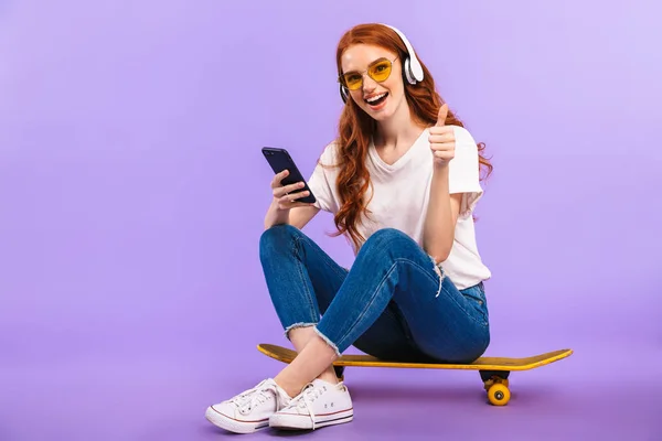 親指を現して ヘッドフォンで音楽を聴く携帯電話を保持している 紫色の背景に分離されたスケート ボードの上に座ってサングラスのうれしそうな若い女の子の完全な長さの肖像画 — ストック写真