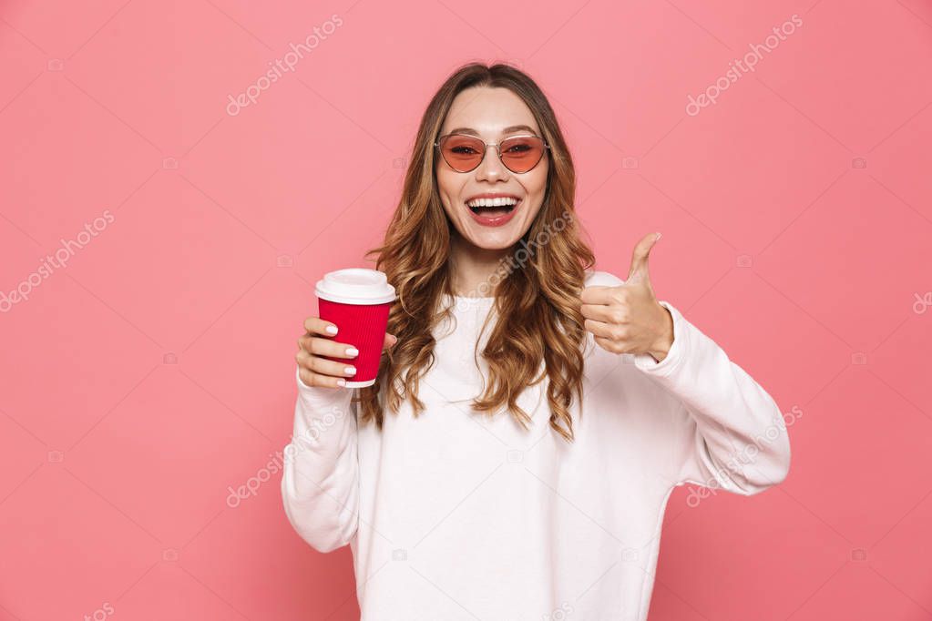 Portrait of a cheerful young woman in sunglasses holding take away coffee isolated over pink background, showing thumbs up