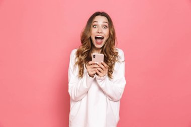 Cep telefonuyla pembe arka plan üzerinde izole heyecanlı bir genç kadın portresi