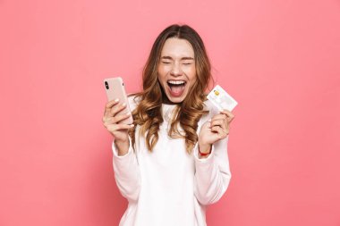 Plastik kredi kartı üzerinde pembe arka plan izole tutan cep telefonuyla bir heyecanlı genç kadın portresi