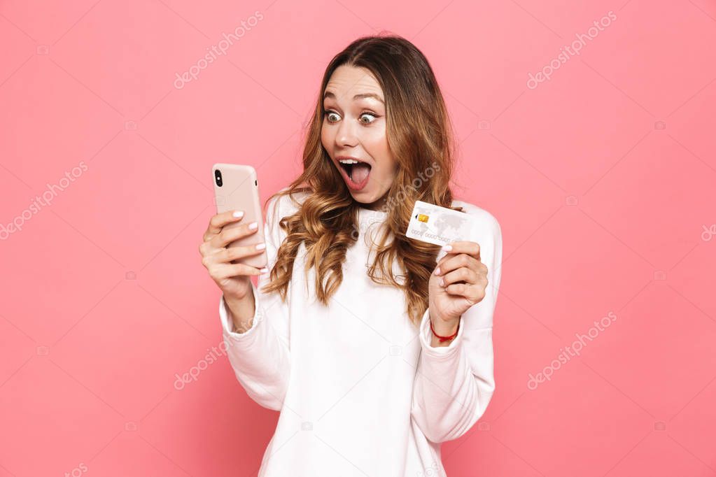 Portrait of a cheerful young woman using mobile phone, holding plastic credit card isolated over pink background