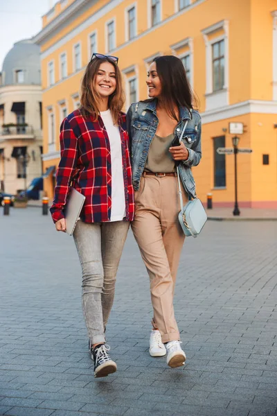 Dos Jóvenes Amigas Sonrientes Caminando Juntas Calle Ciudad Sosteniendo Portátil —  Fotos de Stock