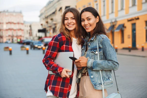 Dos Niñas Sonrientes Amigas Pie Juntas Calle Ciudad Sosteniendo Ordenador —  Fotos de Stock