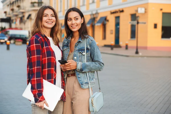 两个快乐的女孩朋友站在一起 在城市的街道 手持笔记本电脑和手机 — 图库照片