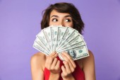 Happy Pretty bruneta žena zakryla tvář s penězi a hledat dál přes fialové pozadí