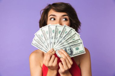 Happy Pretty brunette woman covering her face with money and looking away over purple background clipart