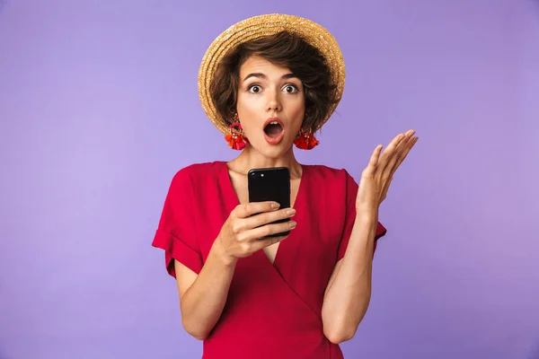 震惊漂亮的黑发妇女在礼服和草帽看着相机 而持有智能手机超过紫色背景 — 图库照片